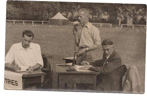 Ernie Goodall standing at a microphone on a sports field in the 1930s, two men seated at a table beside him